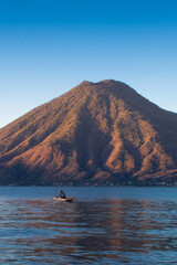 Obraz premium man sailing in a wooden boat on a lake in the middle of a beautiful sunrise with a dormant volcano in the background