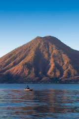 man sailing in a wooden boat on a lake in the middle of a beautiful sunrise with a dormant volcano in the background