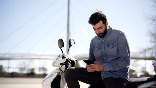Young Spanish man using cell phone or smartphone sitting on his motor scooter outdoors in the city. Businessman checking work issues on cell phone.