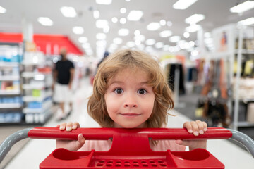 Funny kid with shopping cart buying food at grocery store or supermarket. Surprised kids face.