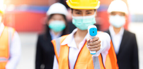 Portrait of Asian woman worker holding thermometer used to measure fever for employees at container yard. worker wearing protection face mask during coronavirus and flu outbreak.