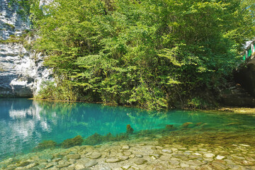 The turquoise water of the mountain lake is transparent. Stones and algae are visible at the bottom. The reservoir is surrounded by white rocks with green vegetation. Blue Lake. Abkhazia