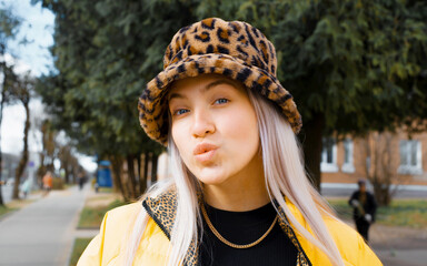 Portrait of young hipster woman in leopard hat making kissing face in camera, outside