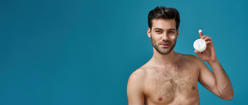 Attractive Naked Man With Brown Hair Holding A White Jar Of Beauty Product, Smiling At Camera, Posing With Cream Applied On His Face Isolated Over Blue Background