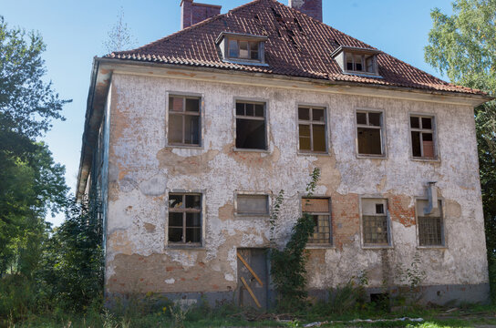 Abandoned dilapidated old brick two-story apartment building with scuffed plaster, broken glass from wooden frames, partially collapsed roof tiles and boarded-up door, with ground overgrown with grass
