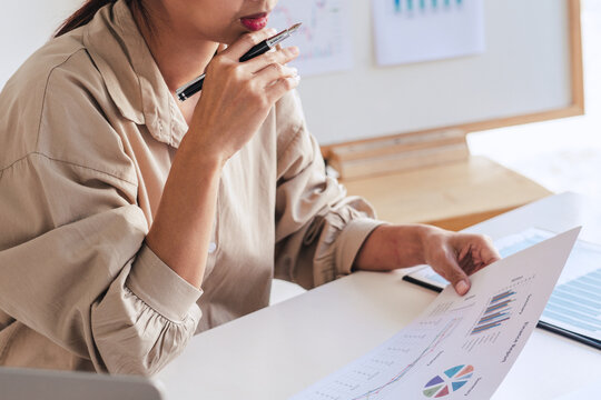 Business, woman holds a pen, look at the paper in the hand, labtop and paper charts on a table, graph board behide