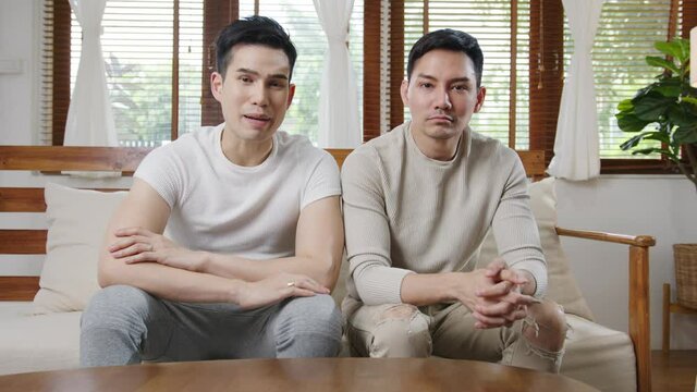 Happy young asian gay couple sit couch use smartphone facetime video call with friends and family in living room at home. Stay at home quarantine, Social distancing, LGBTQ+ Young married concept.