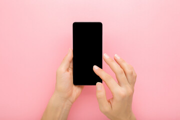 Young adult woman hand holding smartphone and touching black screen with finger on light pink table...