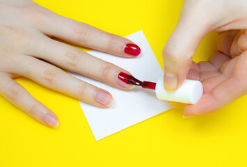 Woman applies red nail polish on the yellow background..Girl making a manicure. Salon procedures at home. Beautiful hands and nails. Close up, macro photo. Colorful and creative.