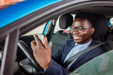 Obraz premium Portrait of young male driver using his mobile phone while driving the car and smiling. Transportation concept - man using phone while driving the car.