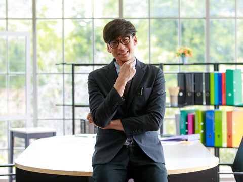 Asian handsome business working man wearing formal black suit, eyeglasses, raising hand to put on chin, large smiling with happiness and success on his job, standing in indoor office or workplace.