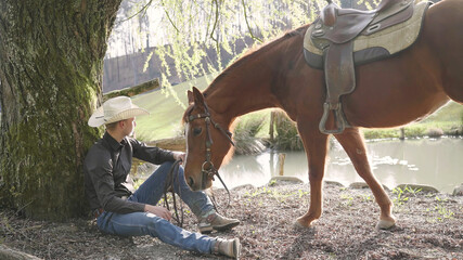Obraz premium Cowboy and horse enjoy time by the lake under a tree