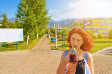 Red-haired woman in dress drinks cocktail on beach. Alley with trees and green lawn. Beautiful girl in striped beach dress is enjoying a summer vacation. Banner for advertising on the background.