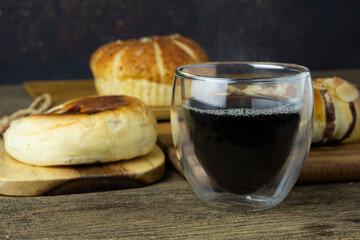 Hot black coffee is placed on the wooden table, there is bread in the background.