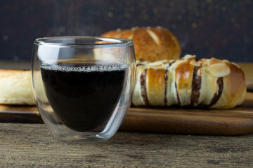 Hot black coffee is placed on the wooden table, there is bread in the background.