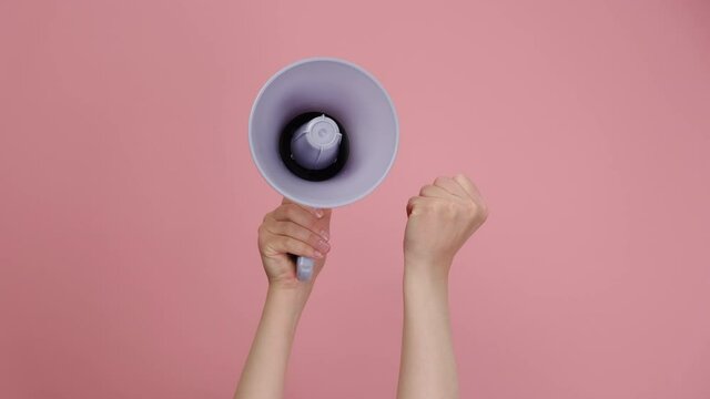Closeup of female hands holding megaphone and raised fist, political protest to showing his power, isolated on pink studio background with copy space for your text or design. Concept revolution, fight