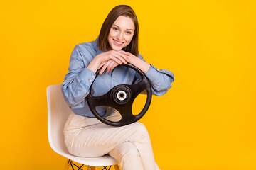 Portrait of attractive cheerful girl sitting on office chair holding steering wheel isolated over bright yellow color background