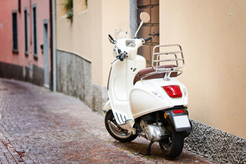 White retro scooter or moped parked on sidewalk of empty narrow street. Motorcycle or motorbike on italian european street