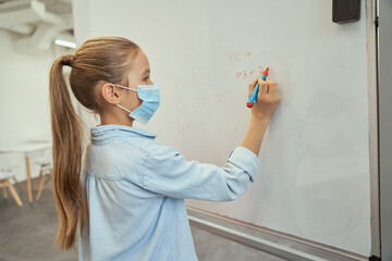 Intelligent little schoolgirl wearing protective mask during coronavirus pandemic writing numbers on board in elementary school classroom