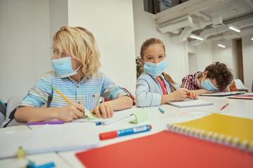 Naklejka premium Little girl wearing protective face mask looking at camera. Elementary school kids sitting together at the table in a classroom