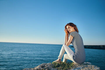 woman in a striped T-shirt and in white trousers in the summer on a stone near the sea in the mountains