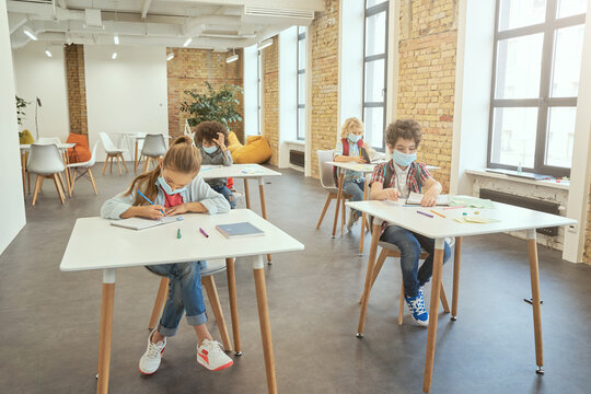 Studying during pandemic. School girl wearing face mask writing in her notebook while sitting at the desk in a classroom. Group of kids in elementary school