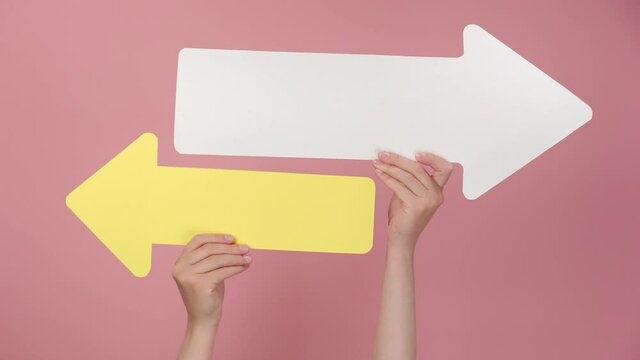 Close up of female hands hold two paper arrow pointing left and right, isolated on pink studio background with copy space for your text or design. Business vision, choice, achievement, target concept