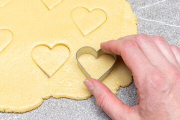 Pastry chef's hand cuts heart-shaped cookies from raw yellow dough on a gray table background, close-up.