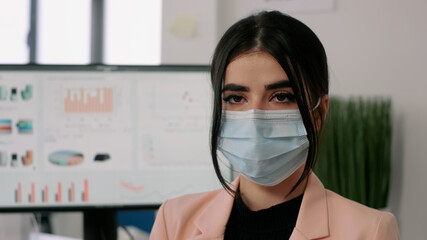 Closeup of businesswoman wearing medical face mask to avoid virus infection during coronavirus lockdown. Entrepreneur looking into camerea while sitting in startup company office.