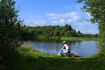 Fototapeta na wymiar thoughtful young girl in a white T-shirt, jeans and a cap on the river bank in summer in sunny weather enjoys the view of the river. Time for solitude and meditation