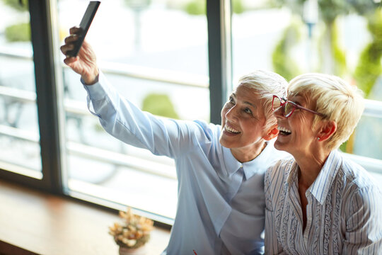 Two Older Business Women Of Similar Appearance Have A Good Time Taking Selfies During A Break At Workplace. Business, Office, Job