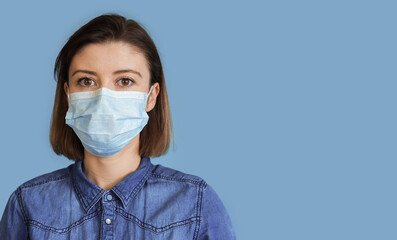 Close up photo of a brunette woman wearing a medical mask on a blue studio wall with free space