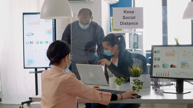 Team workers with protection face masks working in business office on computer during coronavirus lockdown. Coworkers keeping ing ing social distancing to avoid infection with virus disease