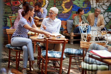Fototapeta na wymiar An older woman and her young female friend talking about a book while they have a drink in the bar. Leisure, bar, friendship, outdoor