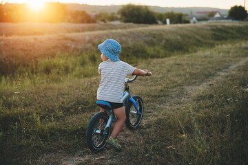 back view photo of a caucasian kid driving the bike in a green field wearing a blue hat