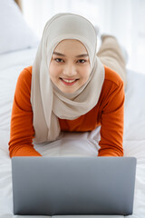 A cute and beautiful Asian Muslim in hijab laying on bed and using tablet notebook computer