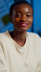 Close up portrait of business african woman smiling at camera after raising head from laptop sitting in startup company late at night. Focused employee doing overtime respecting deadline of project