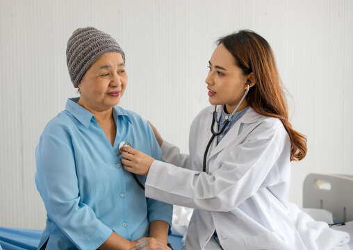 Young And Beautiful Doctor Using Stethoscope For Diagnosis With Older Asian Woman Patient Covered The Head With Clothes Effect From Chemo Treatment In Cancer Cure Process