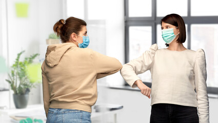 Fototapeta premium health protection, safety and pandemic concept - young women in masks meeting and making elbow bump greeting gesture over office background