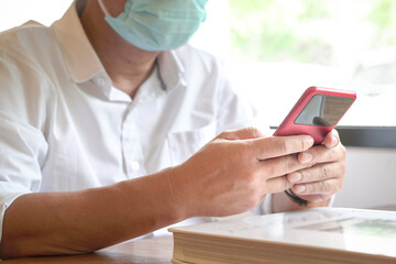 Man with face mask sitting next to windows texting on his phone.