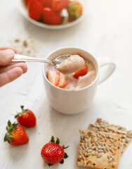 strawberry cocktail in a white cup on a white wooden table decorated with strawberries and cookies. A hand with a spoon, scooping up a smoothie cup. Healthy and Vegetarian Breakfast Concept