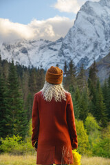 Fototapeta na wymiar View from the back. A blonde woman in a red polto and a brown hat stands in the forest in the mountains against the background of the evening sky and snow-capped mountain peaks
