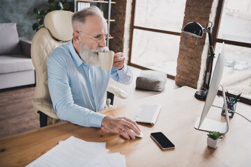 Photo of handsome attentive bearded age agent wear shirt drinking coffee working device indoors workshop workstation workplace