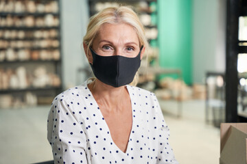Safety. Close up portrait of beautiful mature woman, small business owner wearing protective mask looking at camera while posing in her craft pottery shop