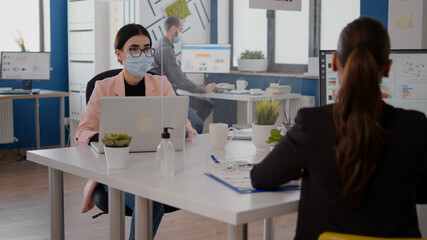 Naklejka premium Freelancer talking with coworker about business startegy while sitting in new normal office wearing protective face mask to prevent infection with coronavirus. Team respecting social distancing