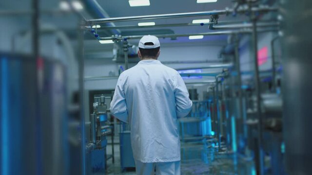 Back rear view of male worker dressed on white uniform standing or walking on modern industrial factory . Man using tablet and doing notes inside milk or juice factory with metal tanks . Slow motion