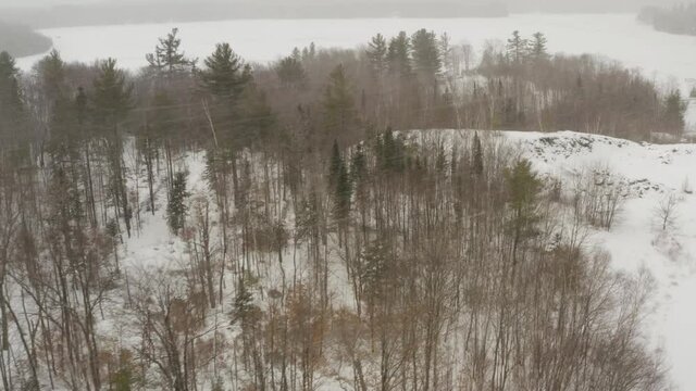 Aerial flight over woods during winter snowfall towards frozen lake  