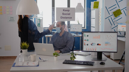 Adult woman with medical face mask measure team temperature using infrared thermometer. Coworkers respecting social distance in new normal company office to avoid infection with virus disease