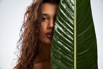 Close up of beautiful female model standing and looking at camera isolated on white. Portrait of young naked woman with perfect makeup posing with mouth open and hiding half of face behind green leaf. - Powered by Adobe