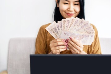happy Asian business woman hand holding Thai baht banknotes setting on sofa , successful with make money online from home concept